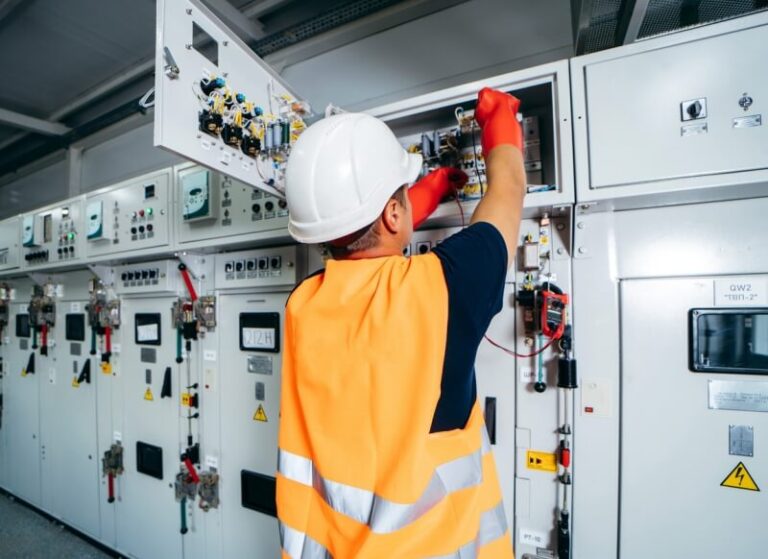 Engenheiro elétrico manuseia grande máquina branca. Ele está de costas, com capacete branco, colete laranja com fitas luminosas e luvas laranjas de borracha